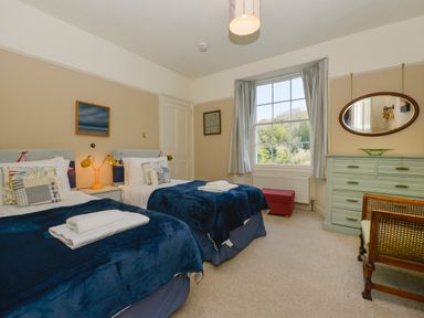 A bedroom with two single beds with blue blankets a window a green dresser a red bench and a wooden chair at Woodlands in Dartmouth