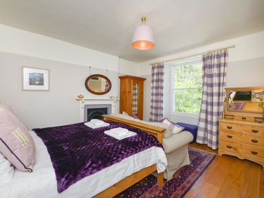 A bedroom with a bed covered with purple blanket two folded towels and pillows a wooden dresser a wardrobe an armchair and a window with plaid curtains at Woodlands in Dartmouth
