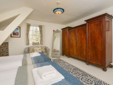A bedroom with two single beds with folded towels a large wooden wardrobe a gray armchair under a window a full length mirror and a blue patterned rug at Woodlands in Dartmouth