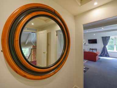 A round mirror on a wall reflecting a hallway with an open door next to a living room with a red sofa a TV and a window with blue curtains at Woodlands in Dartmouth