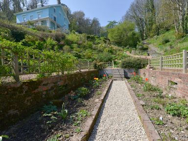 A garden with flower beds along a gravel path and a blue house on a hill at Woodlands in Dartmouth