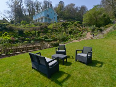 Outdoor seating arrangement on a green lawn with a blue house and garden on a hillside in the background at Woodlands in Dartmouth