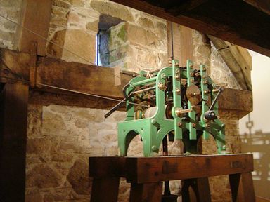 A mechanical clock mechanism mounted on a wooden stand inside a stone wall room at The Clock House in Ivybridge