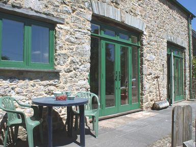 An outdoor patio with a round table and chairs against a stone building with green framed windows and doors at The Clock House in Ivybridge