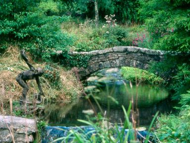 A stone arch bridge over a stream with a bronze statue of a woman beside it in a garden at The Clock House in Ivybridge
