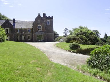 A large stone building with multiple windows and a tower beside a curved gravel driveway surrounded by green lawn and bushes at The Clock House in Ivybridge