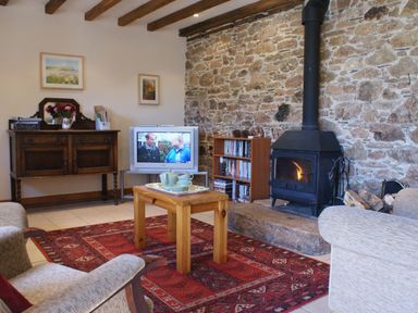 A living room with a wood stove and stone wall a wooden coffee table with a tea set a red patterned rug a television on a stand a wooden chest a bookshelf and exposed ceiling beams at The Clock House in Ivybridge