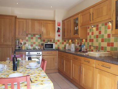 A kitchen with wooden cabinets a tiled multicolored backsplash a table with a yellow floral tablecloth set with plates and glasses at The Clock House in Ivybridge