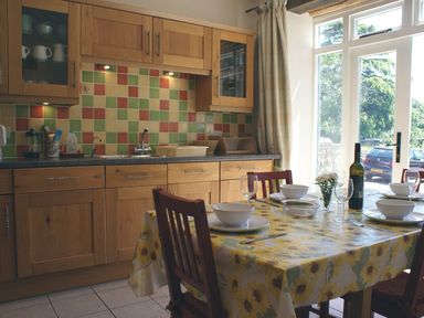 A kitchen with wooden cabinets a tiled backsplash and a dining table set with bowls glasses and a wine bottle near a glass door at The Clock House in Ivybridge