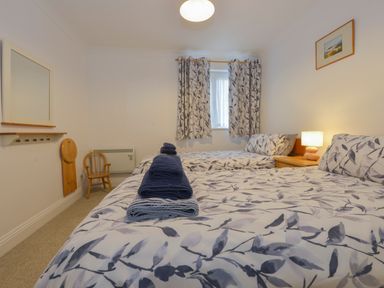 A bedroom with two beds with floral bedding blue towels on one bed a wooden chair a nightstand with a lamp and a window with floral curtains at 5 Burgh Island Causeway in Bigbury-on-Sea