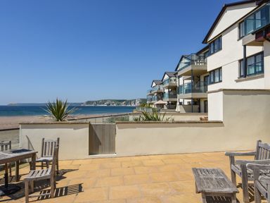A patio with wooden chairs and tables overlooking the beach and sea with modern buildings on the right at 13 Burgh Island Causeway in Bigbury-on-Sea