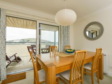 A dining area with a wooden table and chairs next to glass sliding doors leading to a balcony with outdoor furniture at 13 Burgh Island Causeway in Bigbury-on-Sea