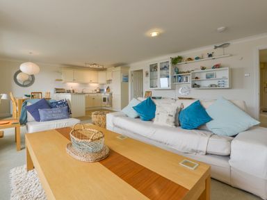 A living room with a sofa and cushions a wooden coffee table and an open kitchen at 13 Burgh Island Causeway in Bigbury-on-Sea