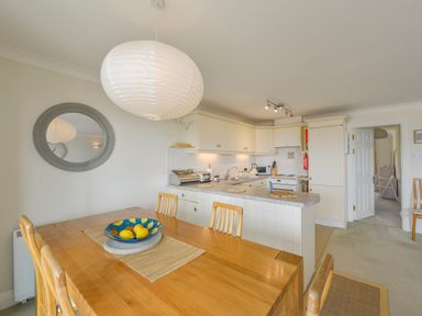 A dining area with a wooden table and chairs next to a kitchen with white cabinets and a countertop at 13 Burgh Island Causeway in Bigbury-on-Sea