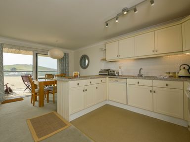 A kitchen and dining area with cream cabinets wooden dining table and chairs and a balcony overlooking a sandy shore at 13 Burgh Island Causeway in Bigbury-on-Sea