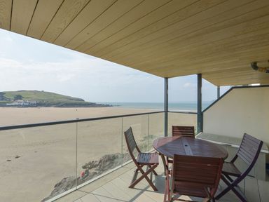 A balcony with a wooden table and four chairs overlooking a sandy beach and ocean at 13 Burgh Island Causeway in Bigbury-on-Sea