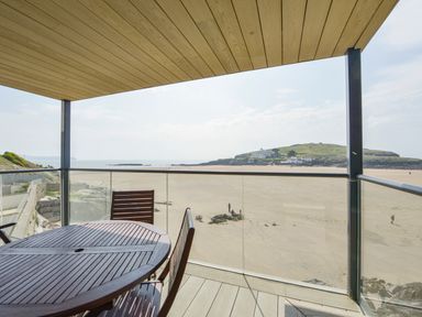 A balcony with wooden table and chairs overlooking a sandy beach and an island with buildings at 13 Burgh Island Causeway Bigbury-on-Sea