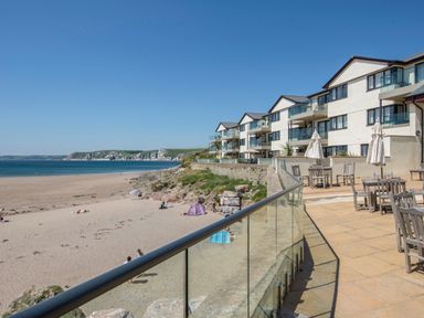 A beachfront view with a sandy beach and a row of residential buildings with balconies and outdoor seating at 13 Burgh Island Causeway in Bigbury-on-Sea