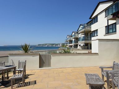 A patio with wooden chairs and tables overlooking the beach and sea with residential buildings beside it at 27 Burgh Island Causeway in Bigbury-on-Sea
