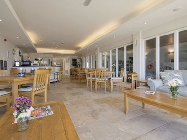A dining area with wooden tables and chairs a sofa and flowers on tables at 27 Burgh Island Causeway in Bigbury-on-Sea