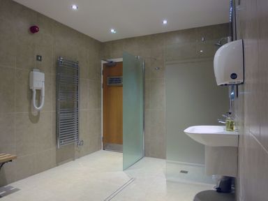 A tiled bathroom with a sink, hand dryer, towel rail, shower area with frosted glass partition, and wooden door at 27 Burgh Island Causeway Bigbury-on-Sea