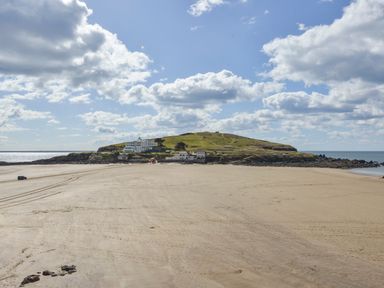 A sandy beach leading to a grass-covered island with buildings under a partly cloudy sky at 24 Burgh Island Causeway in Bigbury-on-Sea