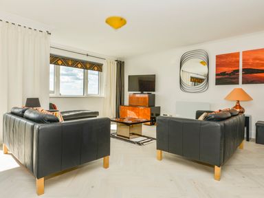 A living room with black leather sofas a wall mirror a television on a wooden cabinet a table and orange paintings at 24 Burgh Island Causeway in Bigbury-on-Sea