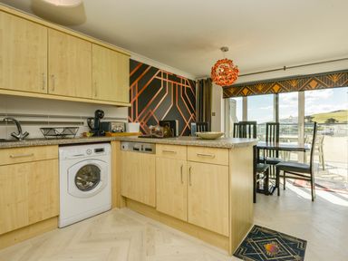 A kitchen with wooden cabinets dishwasher and washing machine next to a dining area with black chairs and large windows at 24 Burgh Island Causeway in Bigbury-on-Sea