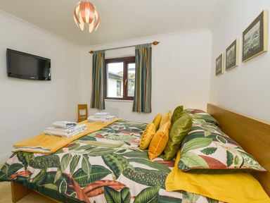 A bedroom with a bed covered in a leaf patterned bedspread and pillows next to a window with striped curtains and a wall-mounted television at 24 Burgh Island Causeway in Bigbury-on-Sea