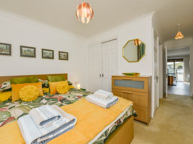 A bedroom with a bed featuring floral bedding and yellow cushions a wooden chest of drawers with a mirror above and a hallway leading to another room at 24 Burgh Island Causeway in Bigbury-on-Sea
