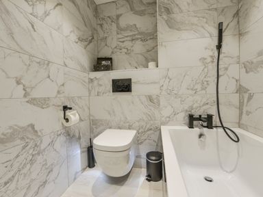 A bathroom with a wall-mounted toilet bathtub with a black faucet and hand shower at 24 Burgh Island Causeway in Bigbury-on-Sea