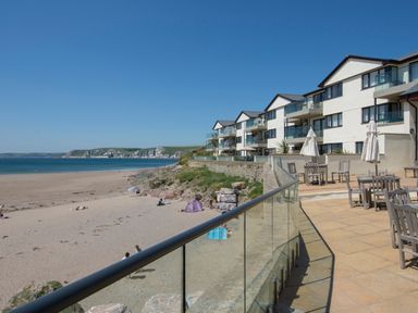 A beachfront patio with wooden tables and chairs next to a modern apartment building overlooking a sandy beach and ocean at 23 Burgh Island Causeway in Bigbury-on-Sea