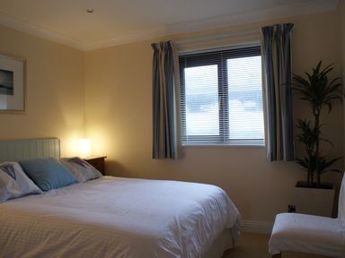 A bedroom with a bed a bedside table with lamp a window with blinds and curtains a chair and a plant at 23 Burgh Island Causeway in Bigbury-on-Sea