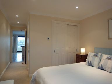 A bedroom with a bed, pillows, a wooden bedside table with a lamp, and a hallway leading to another room at 23 Burgh Island Causeway in Bigbury-on-Sea