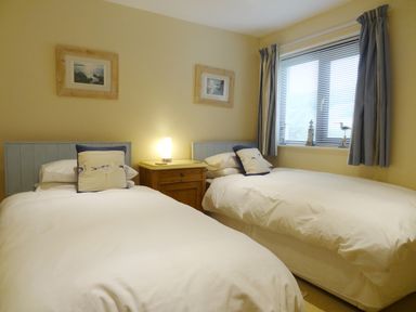 A bedroom with two single beds a wooden nightstand with a lamp a window with curtains and blinds and two framed pictures on the wall at 23 Burgh Island Causeway in Bigbury-on-Sea