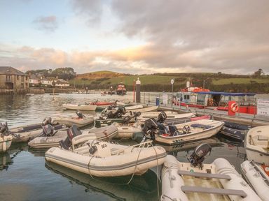 Several small motorboats docked in a harbor with buildings and green hills in the background at Orchard View in Kellaton near Hallsands