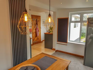 A dining area with hanging pendant lights wooden table and chairs near a window and kitchen at Orchard View in Kellaton near Hallsands