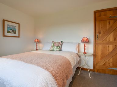 A bedroom with a bed with white bedding a pink blanket and floral pillow two bedside tables with lamps a wooden door and a framed picture on the wall at Orchard View in Kellaton near Hallsands