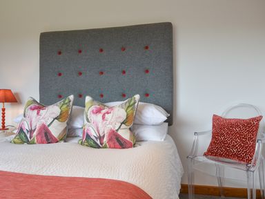 A bed with a gray headboard and floral pillows beside a lamp and a clear chair with a red cushion at Orchard View in Kellaton near Hallsands