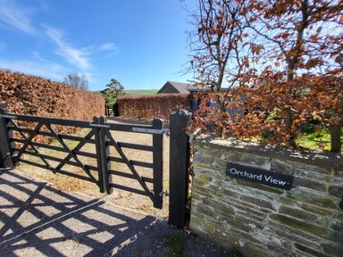 A black wooden gate at the entrance to a gravel driveway with hedges on both sides at Orchard View in Kellaton near Hallsands