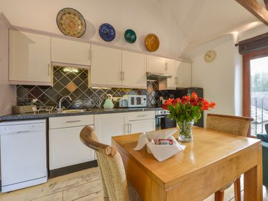 A kitchen with white cabinets black tile backsplash a wooden table with flowers in a vase and woven chairs at Mixit Cottage Kingsbridge