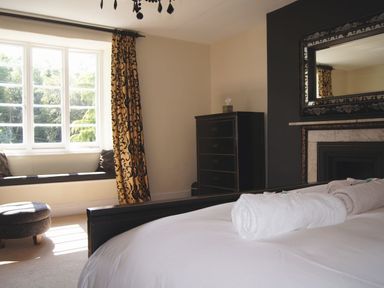 A bedroom with a bed, rolled towels, a fireplace with a mirror, a chest of drawers, a window seat, and patterned curtains at Maristow Barton in Milton Combe near Lopwell Dam