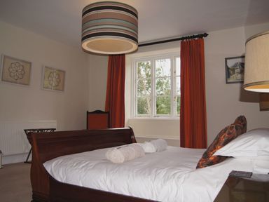 A bedroom with a wooden bed and rolled towels on it a window with red curtains and a striped ceiling light at Maristow Barton in Milton Combe near Lopwell Dam
