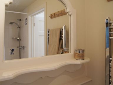 A bathroom mirror reflecting a shower area with a towel on a heated rail and a small jar on a white shelf at Cherry Tree Cottage in Noss Mayo