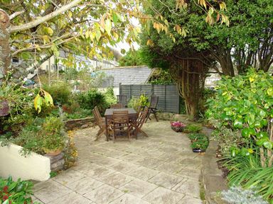 An outdoor patio with wooden table and chairs surrounded by trees and plants at Cherry Tree Cottage in Noss Mayo