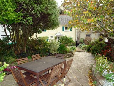 A garden with a wooden table and six chairs on a paved patio surrounded by trees and plants at Cherry Tree Cottage in Noss Mayo