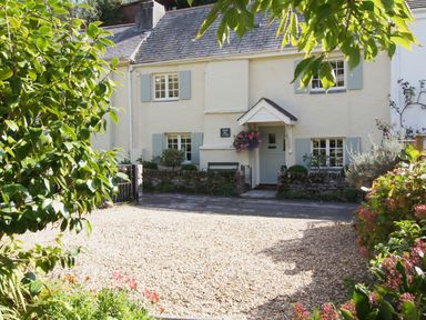 A cottage with light blue shutters and door with hanging flowers and a gravel driveway at Cherry Tree Cottage in Noss Mayo