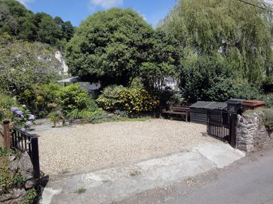 A gravel driveway with black metal gates surrounded by trees and shrubs at Cherry Tree Cottage in Noss Mayo