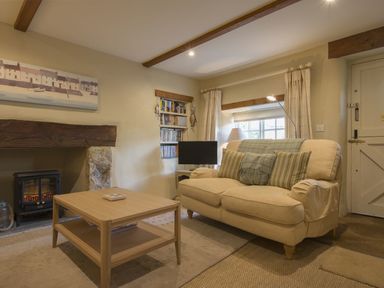 A living room with a beige sofa pillows a wooden coffee table a small fireplace a bookshelf and a window at Cherry Tree Cottage in Noss Mayo