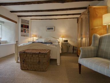 A bedroom with a bed two bedside tables with lamps a wicker chest an armchair and a wooden wardrobe at Cherry Tree Cottage in Noss Mayo
